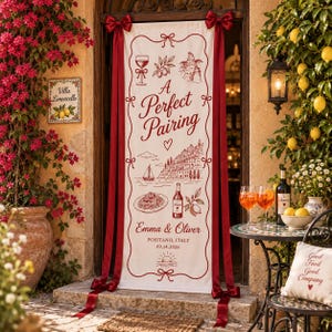 May include: A vertical cream-colored fabric banner with dark red ink illustrations and text reading A Perfect Pairing, Emma & Oliver, Positano, Italy, 09.14.2026. The banner features a wine glass, pasta, a bottle, and a coastal scene, tied with red ribbons.