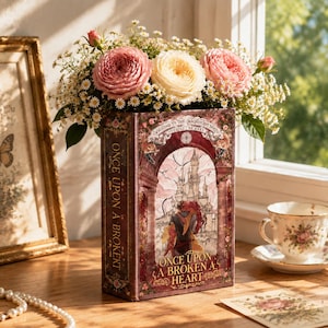 Pode incluir: Um vaso retangular em forma de livro em tons de vermelho profundo e dourado, com uma ilustração de um castelo e duas figuras. O texto frontal diz "Once Upon a Broken Heart" e "Welcome to the Magnificent North". Está cheio de rosas cor-de-rosa e brancas.