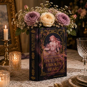 May include: A hardcover book titled Once Upon a Broken Heart by Stephanie Garber with a dark purple cover featuring gold filigree, an illustration of a figure with long hair, and the text Grace Zhu Art, topped with a bouquet of purple and cream roses.
