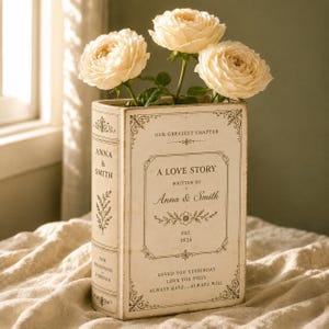 May include: A cream-colored ceramic vase shaped like a book, featuring the text "A LOVE STORY," "ANNA & SMITH," "EST. 2026," and "OUR GREATEST CHAPTER." Three pale cream roses are arranged inside the vase, which has decorative black filigree borders.