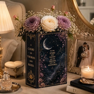 Pode incluir: Um livro decorativo intitulado A Court of Thorns and Roses com uma capa azul-escura apresentando uma lua crescente, um castelo e o texto To the stars who listen and the dreams that are answered, coberto por um buquê de rosas roxas e brancas.
