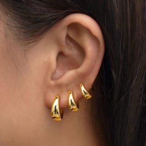 Peut inclure: Trois boucles d'oreilles dorées de tailles différentes sont portées sur une oreille. Les boucles d'oreilles ont un design lisse et incurvé, reflétant la lumière. La plus grande boucle d'oreille est la plus proche du lobe, avec deux plus petites remontant l'oreille.