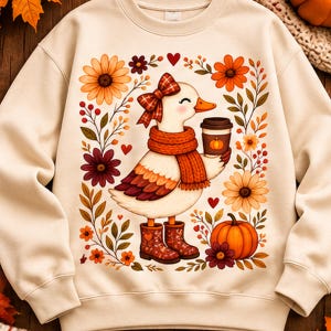 Peut inclure: Un sweat-shirt à col rond couleur crème avec un motif central représentant un canard blanc portant un nœud à carreaux rouges, une écharpe orange et des bottes à motifs. Le canard tient un gobelet de café, entouré de citrouilles et fleurs.