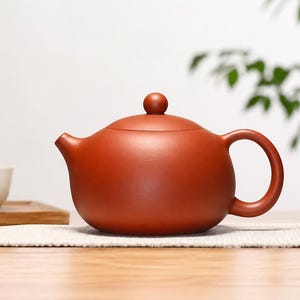 Peut inclure: Une théière en céramique brun-rouge avec un corps arrondi, une anse incurvée et un petit bec verseur. Le couvercle est orné d'un petit bouton rond. La théière est posée sur un tissu blanc, avec une tasse à thé et un plateau en bois en arrière-plan.