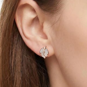 Peut inclure: Boucles d'oreilles à clous en forme de feuille en or rose avec des cristaux transparents. Les boucles d'oreilles ont la forme d'une feuille avec une tige et sont serties de petits cristaux étincelants.
