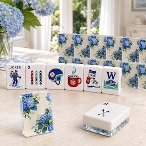 May include: A set of white mahjong tiles featuring blue hydrangea floral patterns on the backs. The front faces display colorful winter-themed illustrations including a skier, skis, a helmet, a mug of cocoa, a ski boot, a chalet, and the number 8.