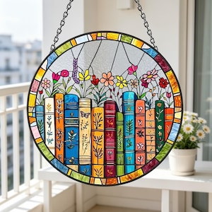 May include: A round stained glass window featuring a colorful design of books and flowers. The books are various colors, and the flowers include pink, yellow, and red blooms. The window is framed in black and hangs from a chain.