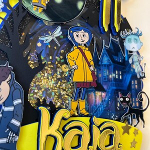 May include: A layered paper art piece featuring characters from a fantasy movie. The design includes a girl in a yellow raincoat, a black cat, and a haunted house. The name "Kaja" is prominently displayed in gold lettering.