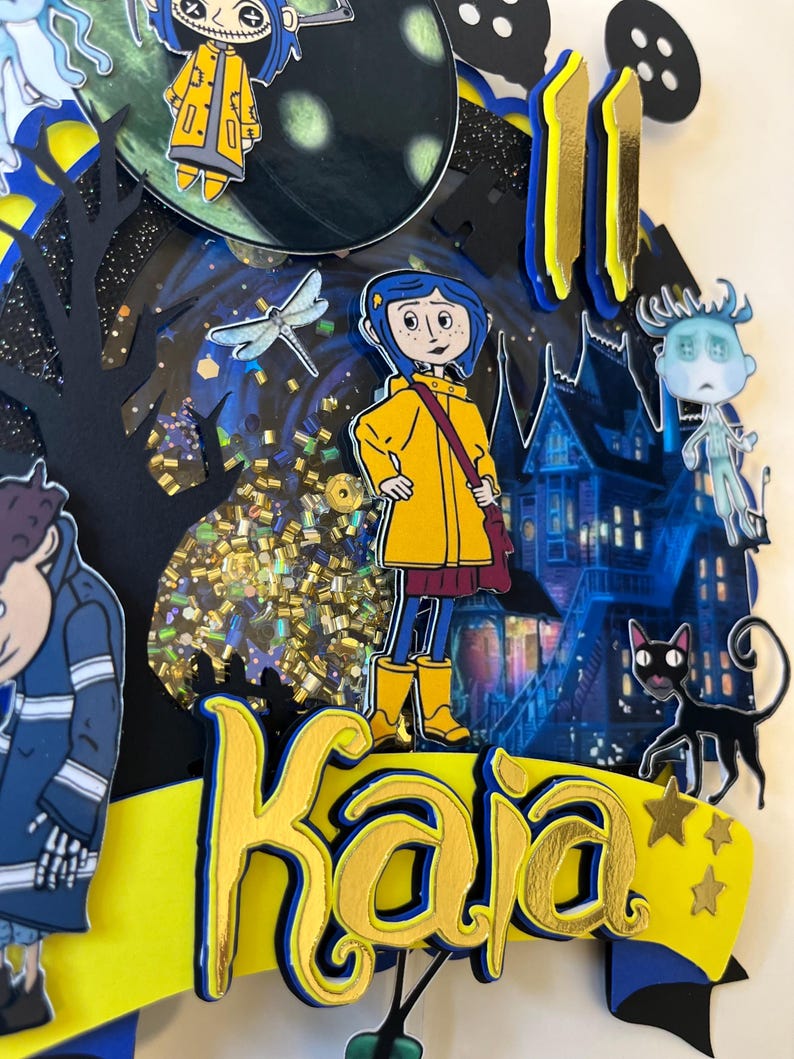 May include: A layered paper art piece featuring characters from a fantasy movie. The design includes a girl in a yellow raincoat, a black cat, and a haunted house. The name "Kaja" is prominently displayed in gold lettering.