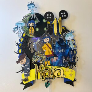 May include: A layered cake topper featuring characters from the animated film, "Coraline." The design includes a yellow banner with the name "Kaia" in gold lettering, a black cat, and a haunted house. The number "11" is also included.