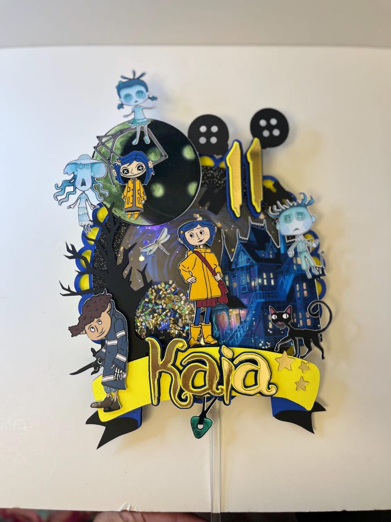 May include: A layered cake topper featuring characters from the animated film, "Coraline." The design includes a yellow banner with the name "Kaia" in gold lettering, a black cat, and a haunted house. The number "11" is also included.