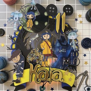 May include: A layered cake topper featuring characters from the animated film "Coraline." The design includes a yellow banner with the name "Kaia," a black cat, and a spooky house. The topper is surrounded by macarons and star-shaped confetti.
