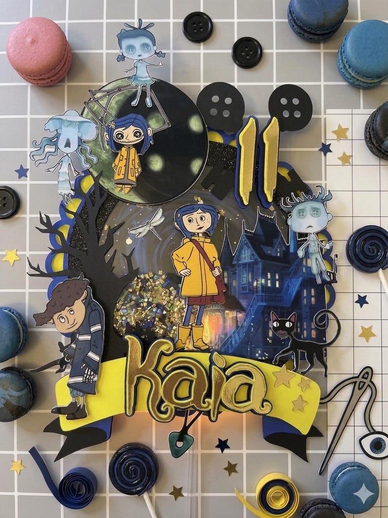 May include: A layered cake topper featuring characters from the animated film "Coraline." The design includes a yellow banner with the name "Kaia," a black cat, and a spooky house. The topper is surrounded by macarons and star-shaped confetti.