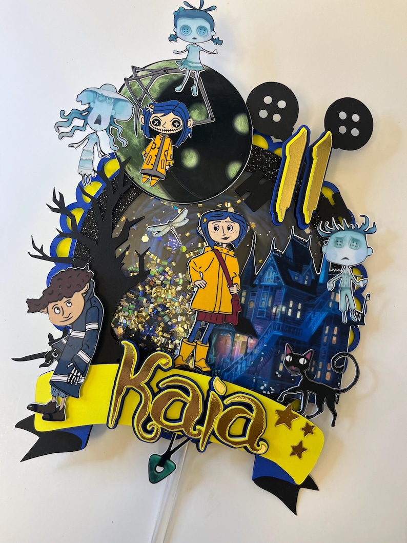 May include: A layered cake topper featuring characters from the animated film "Coraline." The design includes a yellow banner with the name "Kaja," a haunted house, black cat, and various character cutouts. The number "11" is also included.