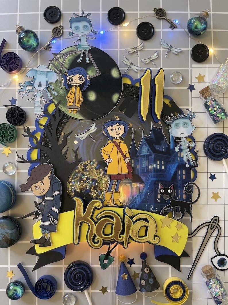 May include: A colorful cake topper featuring characters from the animated film "Coraline." The design includes a central image of the character, the name "Kaia," and the number "11." Decorative elements include buttons, keys, and miniature bottles.