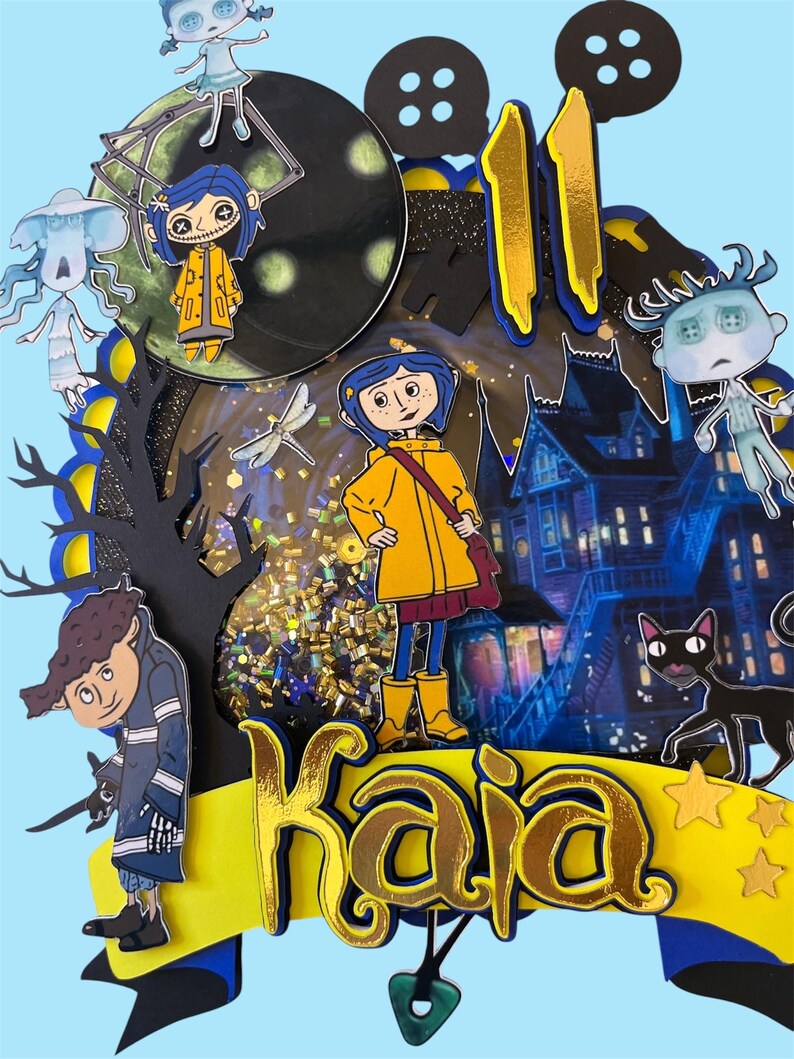 May include: A layered, colorful cake topper featuring characters and elements from the animated film "Coraline." The design includes the name "Kaia" in gold lettering, the number "11," and various character cutouts, all set against a blue background.