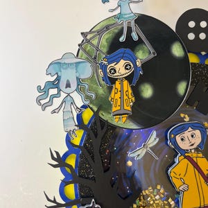 May include: A colorful, layered artwork featuring characters from the animated film "Coraline." The design includes cut-out figures, a dark tree silhouette, a large button, and a glittery, textured element. The color palette includes blue, yellow, and black.