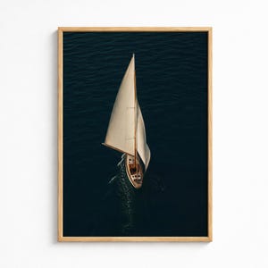 May include: A framed vertical wall art print featuring an aerial view of a single sailboat with white sails navigating dark blue water. The boat has a wooden deck and hull, creating a wake as it moves through the deep sea, enclosed in a light wood frame.