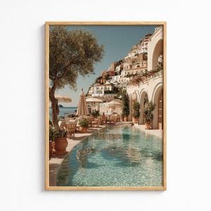 May include: A framed print depicts a scenic coastal view. A clear pool reflects the sky and surrounding architecture. White buildings cascade down a hillside, with a restaurant and umbrellas near the pool. An olive tree and potted plants add to the ambiance.