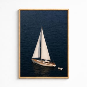 Puede incluir: Una impresión de arte mural enmarcada que muestra una vista aérea de un velero blanco con dos velas navegando en aguas oceánicas azul profundo. Un pequeño bote auxiliar blanco está atado a la popa del barco. La obra tiene un marco de madera clara.