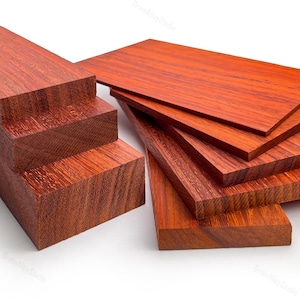 May include: A collection of rectangular wooden blocks and flat boards featuring a deep reddish-brown color with a visible, dense grain pattern. The pieces are arranged in a stacked formation, showcasing the smooth, sanded surfaces and edges.