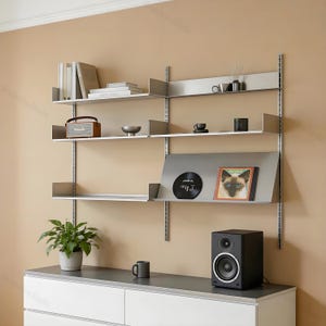 May include: A modular wall-mounted shelving system with silver metal uprights and shelves. The shelves hold books, a brown radio, a small bowl, mugs, a black vinyl record, and a framed cat portrait. A black speaker sits on a white cabinet below.