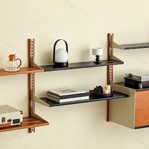 May include: A modular wall-mounted shelving system featuring vertical wooden tracks with metal brackets supporting horizontal black and brown wooden shelves. The shelves hold a glass teapot, a white globe lamp, a white bowl, books, and a record player.