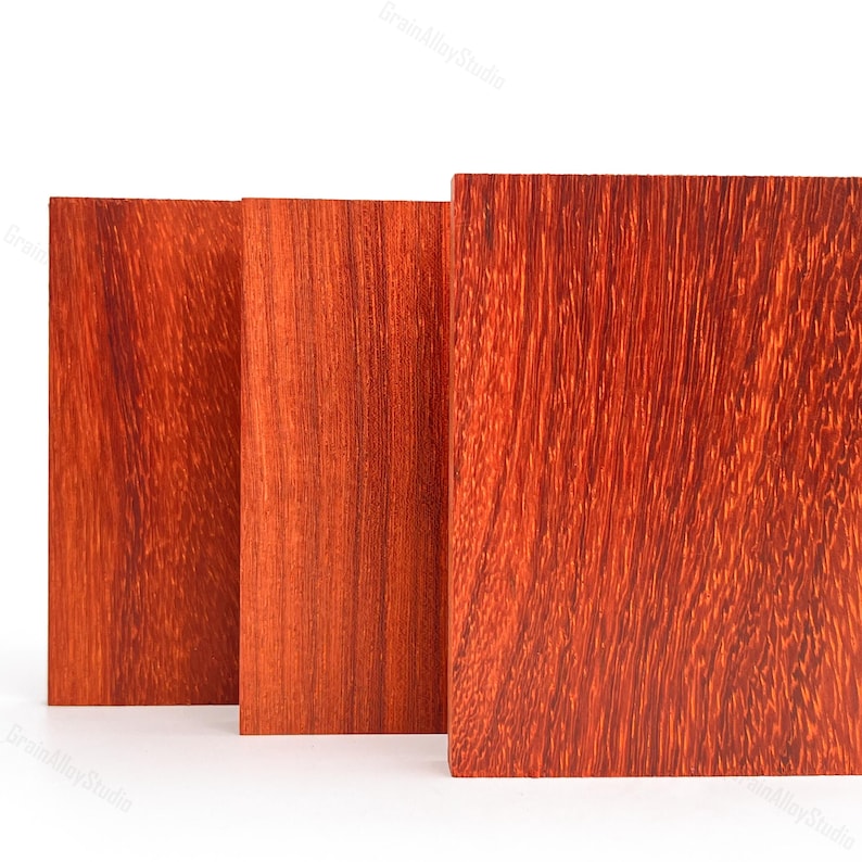 May include: Three rectangular wooden boards standing upright, featuring a vibrant reddish-orange hue with a distinct, dense, and linear grain pattern visible across the smooth, flat surfaces of each piece of lumber.
