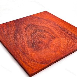 May include: A square wooden board featuring a vibrant reddish-orange hue with a prominent, swirling burl grain pattern. The surface displays intricate, dark concentric rings and organic textures that radiate from a central knot-like formation.