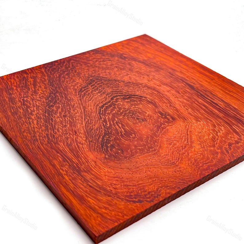May include: A square wooden board featuring a vibrant reddish-orange hue with a prominent, swirling burl grain pattern. The surface displays intricate, dark concentric rings and organic textures that radiate from a central knot-like formation.