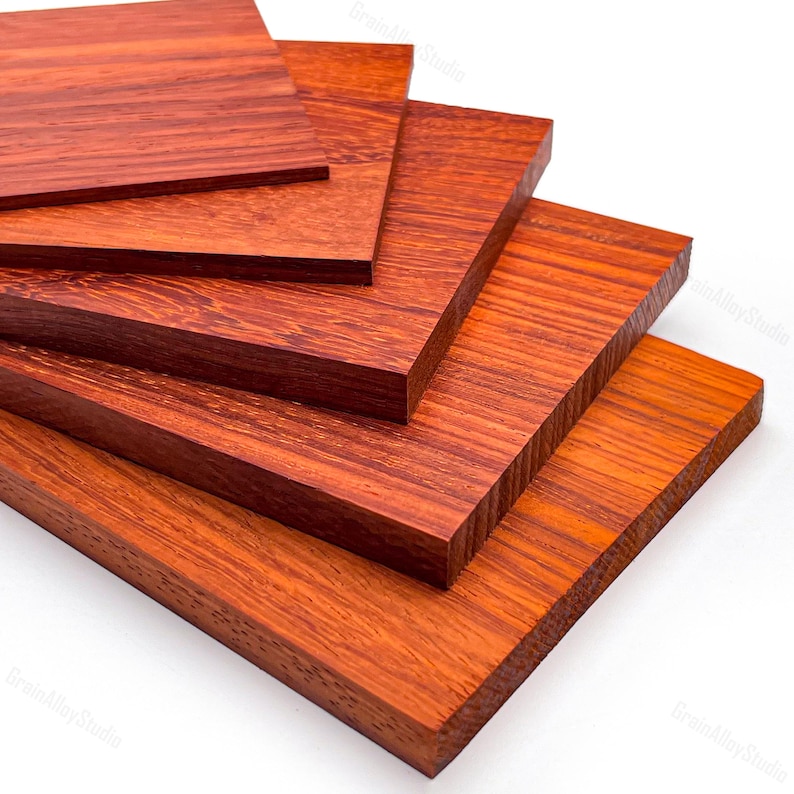 May include: A stack of five rectangular wooden boards arranged in a cascading fan pattern. Each board features a rich, reddish-brown hue with a visible, linear wood grain texture running horizontally across the flat, smooth surfaces.