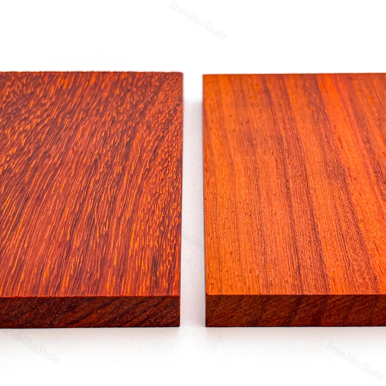 May include: Two rectangular wooden blocks placed side by side. The left block features a deep reddish-brown hue with a dense, textured grain pattern. The right block displays a lighter, vibrant orange-red tone with a straighter, smoother grain.