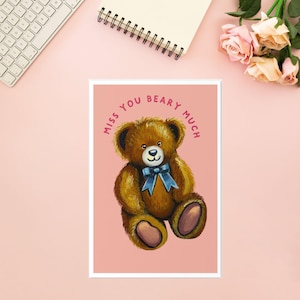 May include: A framed print featuring a brown teddy bear with a blue bow tie, set against a pink background. The text "MISS YOU BEARY MUCH" is arched above the bear. The image is surrounded by a white border.
