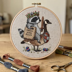 May include: An embroidery hoop featuring a raccoon wearing a green leaf crown, holding a pizza box labelled TRASH KING PIZZA, riding a goose equipped with a wooden sword and a shield. Two soda cans lie on the ground below the goose.