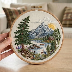 May include: A circular embroidery hoop containing a fabric panel with a detailed landscape scene featuring a large pine tree, snow-capped mountains, a yellow sun, a small cabin, a lake with a boat, and a bird, all rendered in colourful thread.