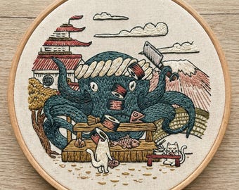 May include: Embroidered artwork in a wooden hoop depicts a blue octopus chef preparing sushi. The scene includes a Japanese-style building, Mount Fuji, and cats. The octopus wears a chef's hat and holds a knife, with sushi rolls nearby.