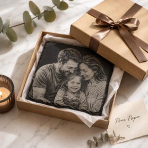 Puede incluir: Una placa cuadrada de pizarra negra grabada con un retrato en escala de grises de una familia compuesta por un hombre, una mujer y un niño. La placa está dentro de una caja de regalo de cartón beige con una cinta de satén marrón y una nota.