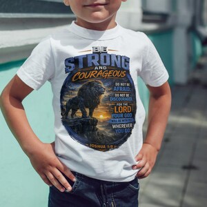 May include: A white short-sleeved t-shirt featuring a circular graphic of a lion standing on a cliff at sunset. The text reads: BE STRONG AND COURAGEOUS, DO NOT BE AFRAID; DO NOT BE DISCOURAGED, FOR THE LORD YOUR GOD WILL BE WITH YOU WHEREVER YOU GO. JOSHUA 1:9.