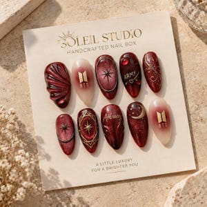 Peut inclure: Un ensemble de dix faux ongles sur une carte de présentation marquée Soleil Studio Handcrafted Nail Box. Les ongles présentent des tons rouge foncé, bordeaux et blanc translucide avec des accents dorés, incluant des logos BTS, des étoiles, des lunes.