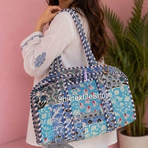 May include: A quilted duffel bag featuring a patchwork design in various shades of blue and white with floral and geometric patterns. The bag has two shoulder straps with a blue and white striped trim and a gold-tone metal buckle. Text reads ShivtextileShop.