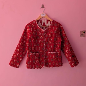 Könnte beinhalten: Rote Steppjacke mit floralem Muster und weißer Bordüre. Die Jacke hat lange Ärmel, zwei Vordertaschen und einen V-Ausschnitt. Sie hängt an einem Holzkleiderbügel vor einer rosa Wand.
