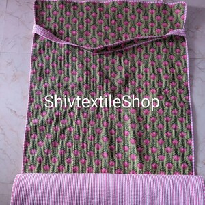 Könnte beinhalten: Ein rechteckiges gestepptes Textil mit einem sich wiederholenden Muster aus rosa Blütenknospen auf grünen Stielen vor einem dunkelgrünen Hintergrund. Die Kanten sind mit rosa-weiß gestreiftem Stoff eingefasst und der untere Teil ist aufgerollt.