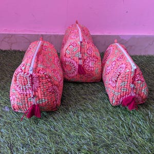 Peut inclure: Trois pochettes en tissu matelassé de couleur corail vif, ornées d'un motif floral détaillé dans des tons de rose, rouge et vert. Chaque pochette est dotée d'une fermeture éclair blanche avec un petit pompon en tissu rouge foncé.