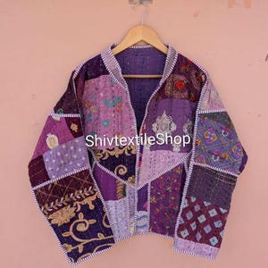 May include: A long-sleeve jacket constructed from various purple and violet fabric patches featuring intricate embroidery, floral patterns, and geometric designs. The garment is finished with visible white running stitch detailing and the text ShivtextileShop.