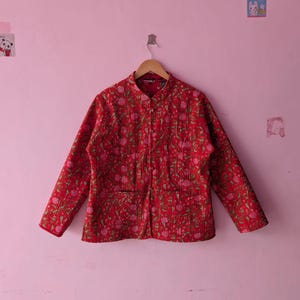 Peut inclure: Une veste matelassée à manches longues d'un rouge vif avec un motif floral répétitif dans des tons de rose et de vert. La veste présente un col officier, une fermeture boutonnée sur le devant et deux poches avant, suspendue sur un cintre en bois.