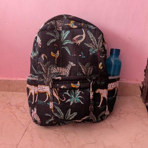 Könnte beinhalten: Ein schwarzer, gesteppter Rucksack mit einem sich wiederholenden Muster aus Zebras, Leoparden und tropischem Blattwerk in Weiß-, Grün- und Gelbtönen. Der Rucksack verfügt über eine Reißverschlusstasche vorne und ein Seitenfach mit einer blauen Wasserflasche.