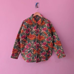 May include: A floral print jacket with a button-down front and two front pockets. The jacket features a colourful pattern of red, orange, and green flowers on a cream background. It hangs on a wooden hanger against a pink wall.