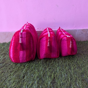 Puede incluir: Tres estuches acolchados de diferentes tamaños, con un vibrante patrón de rayas en rosa intenso y rojo oscuro. Cada estuche está confeccionado con una tela suave y texturizada, una cremallera blanca y una borla roja a juego en el tirador.