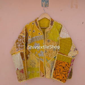 Op de afbeelding: Een geel patchwork jasje met diverse bloem- en geometrische patronen in mosterd-, goud- en wittinten. Het kledingstuk heeft lange mouwen, een opstaande kraag en zichtbare kantha-stiksels. De tekst ShivtextileShop is zichtbaar.