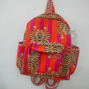 May include: A colourful backpack with a tiger print. The bag has orange and pink vertical stripes, with tiger illustrations. It features a front pocket, side pockets, and adjustable straps. A water bottle is in the side pocket.